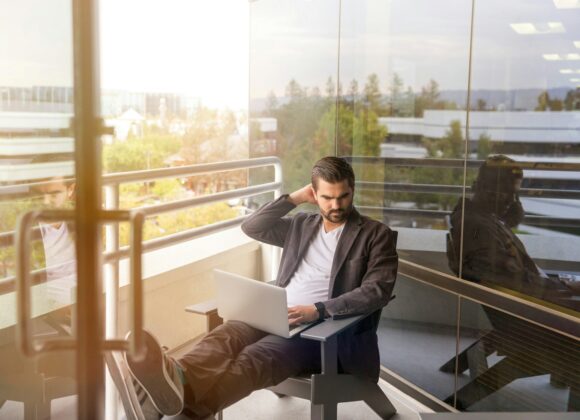 man sitting on gray arm chair using silver laptop computer on building balcony at daytime